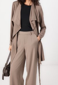 Jasnobrązowy, oversize blazer z drapowanym przodem, zestawiony z spodniami o wysokim stanie i szerokimi nogawkami. Czarna bluzka i ciemna torebka dopełniają cały look.