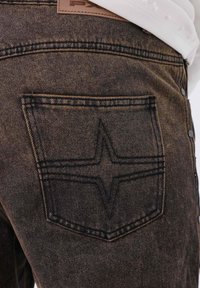 Acercamiento del bolsillo de unos jeans de mezclilla oscuros con costuras en forma de estrella negras y detalles de botones en la costura lateral.