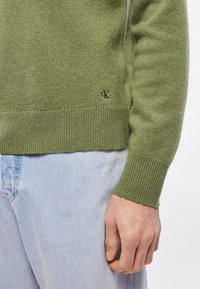 Zöld kötött pulóver bordázott mandzsettákkal és szegéllyel, oldalán egy kis "cK" logóval. Világoskék farmernadrággal párosítva.