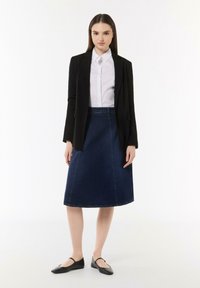 Schwarze taillierte Blazer über einem weißen Kragenhemd, kombiniert mit einem dunklen Jeans-A-Linien-Rock und schwarzen flachen Schuhen, stehend vor einem schlichten Hintergrund.