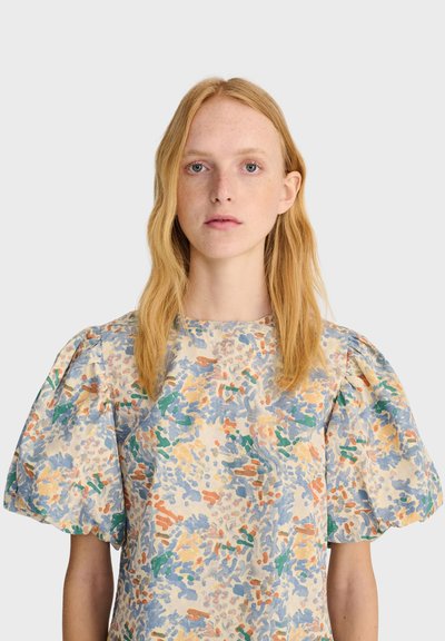 Jeune femme aux cheveux blonds et raides portant un chemisier à manches courtes bouffantes avec des motifs floraux bleus, orange, verts et jaunes.