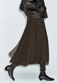 Maxi gonna di un colore marrone scuro e fluida, realizzata in tessuto leggero, abbinata a stivali neri in pelle aderenti fino alla caviglia e a una giacca bomber nera in pelle.