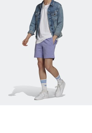 Persona con chaqueta de mezclilla azul claro, camiseta blanca, pantalones cortos lila, zapatillas blancas y calcetines blancos con rayas azules, de pie contra un fondo liso.