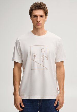 Junger Mann mit lockigem Haar, der ein weißes T-Shirt mit einem minimalistischen Strandmotiv trägt, vor einem einfarbigen hellen Hintergrund stehend.
