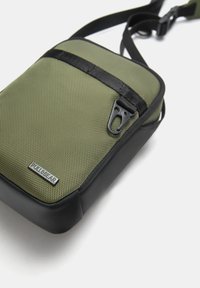 Grøn tekstureret nylon crossbody taske med en sort bund, der har lynlås, justerbar rem og et metal logo label.