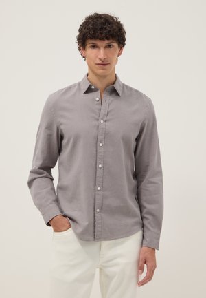 Joven con cabello rizado, vistiendo una camisa gris de manga larga abotonada y pantalones blancos, de pie con una mano en el bolsillo.