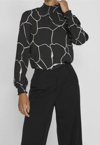 Černá blůza s bílým abstraktním vzorem, smockovaný výstřih, dlouhé rukávy se staženými manžetami, nošena s černými kalhotami. Hladká textura látky.