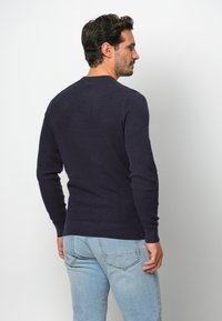 Marineblauer Pullover mit langen Ärmeln, Rundhalsausschnitt, gerippten Bündchen und Saum, getragen mit hellblauen Jeans, die den strukturierten Stoff und das schmale Design zeigen.