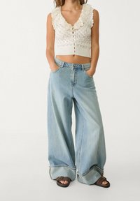 Biały crop top wydziergany na szydełku z falbaniastym dekoltem i zapięciem na guziki, zestawiony z oversize'owymi, jasnoniebieskimi spodniami w palazzo z podwiniętymi mankietami.