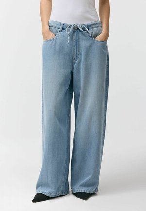 Osoba ubrana w jasnoniebieskie szerokie dżinsy z rękami w przednich kieszeniach oraz czarne buty z ostrym noskiem.