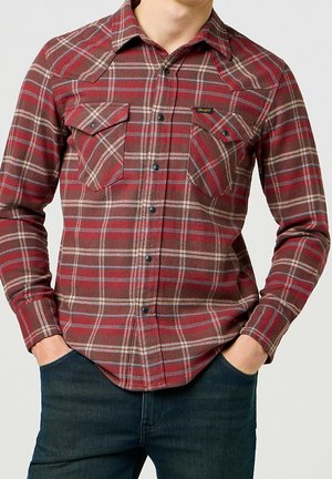 Mand iført rød og brun ternet skjorte med knapper og to brystlommer og mørkeblå jeans, hænderne i lommerne.