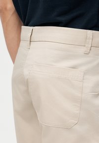 Pantalon beige avec une poche arrière porté avec une chemise noire, montrant les passants de ceinture et un gros plan sur la texture du tissu et les coutures.