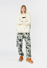 Sudadera beige con detalle de bolsillo negro, combinada con pantalones grises estampados que tienen acentos en verde y negro. Calzado marrón completa el conjunto.