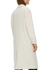 Lång vit kappa i texturerat vävt tyg, med slagkrage och uppvikta ärmslut. Har slits bak och rak silhuett.