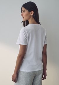 T-shirt bianca in cotone con maniche corte e scollatura rotonda, indossata da un modello di spalle contro un muro chiaro.