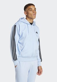 Ljusblå zip-up huvtröja i mjukt tyg, med svarta tre-stripes detaljer på ärmarna och en framstående ficka.