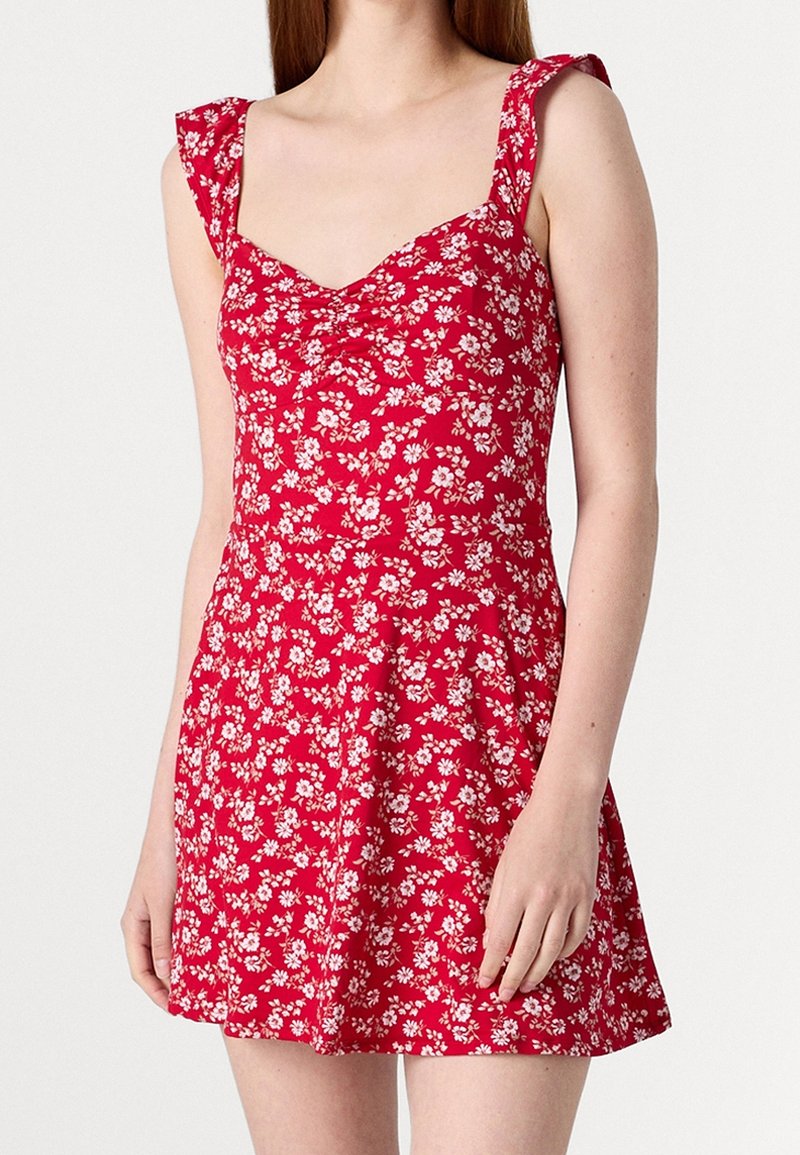 Punainen kukallinen mekko, jossa on istuva yläosa ja levenevä helma. Ominaisuuksiin kuuluvat lyhyet, röyhelöiset olkaimet ja pieniä valkoisia kukkakuvioita kankaassa.