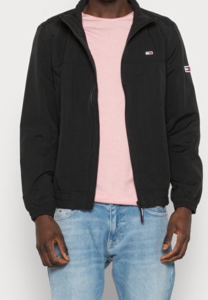 Mand iført sort lynlåstrøje over lyserød t-shirt og lyseblå jeans, stående med hænderne afslappet langs siden.