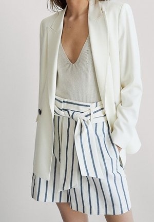 Femme portant un blazer blanc, un haut en maille beige, et un short blanc taille haute à rayures verticales bleues avec une ceinture nouée.