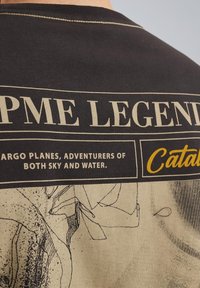 Camiseta negra con diseño gráfico; presenta texto e ilustraciones en color beige. Los detalles clave incluyen la marca “PME Legend” y la imagen de un avión de carga.