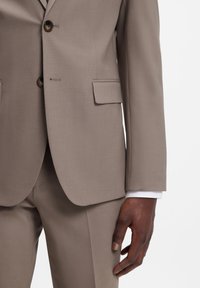 Μπεζ σακάκι ανδρικού κοστουμιού με δύο κουμπιά, πλαϊνή τσέπη με φλάπ και ορατό καφέ χέρι δίπλα του, σετ με παντελόνι ίδιο χρώμα.
