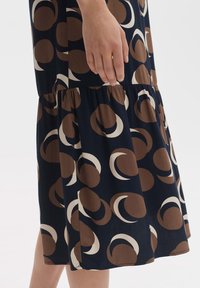 A navy dress featuring a tiered design with a circular brown and cream pattern. Made of soft, lightweight fabric. Closure not visible.