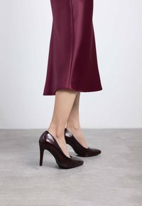 Zapatos de tacón alto en cuero burdeos profundo, con una superficie lisa, punta afilada y forma contorneada, sobre un fondo gris claro.