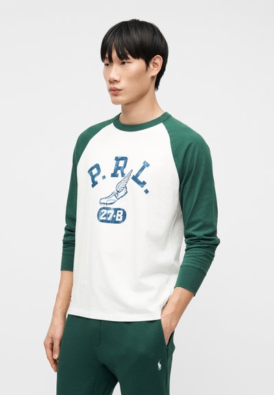 Νεαρός άνδρας που φοράει λευκό και πράσινο μακρυμάνικο πουκάμισο με το λογότυπο "P.R.L." και ένα σχέδιο με φτερωτό παπούτσι, συνδυασμένο με πράσινο παντελόνι με μικρό λευκό λογότυπο.