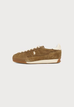 Baskets en daim marron avec lacets blancs, semelle en caoutchouc et logo blanc sur le côté, présentées sur un fond clair uni.