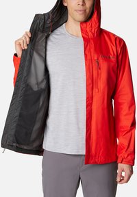 Leichte rot-schwarze Jacke mit Kapuze; verfügt über eine Reißverschlusstasche auf der Brust und ein Mesh-Futter; wird offen über einem grauen T-Shirt getragen.