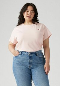 Camiseta de algodón rosa claro con mangas cortas, cuello redondo y pequeño logotipo rojo en el pecho, combinada con jeans de mezclilla azul de cintura alta.