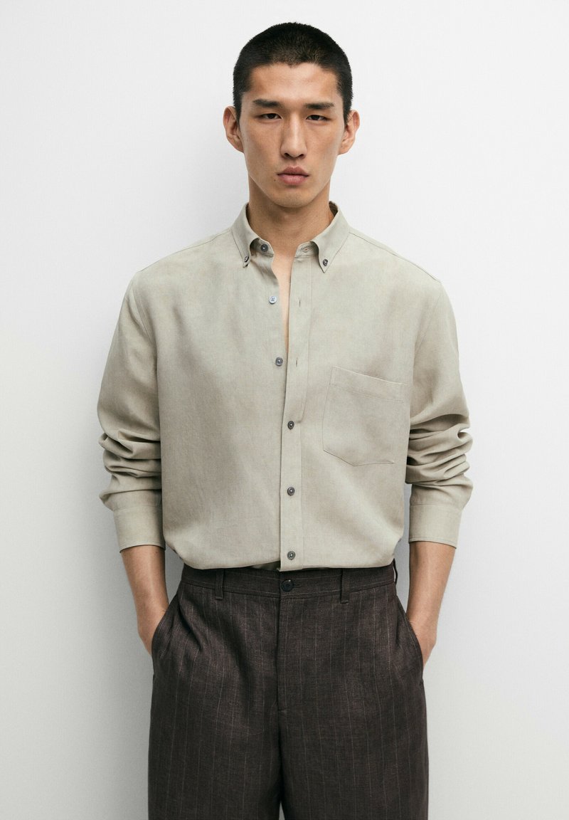 Giovane uomo con capelli corti che indossa una camicia beige chiaro con bottoni e pantaloni scuri a righe sottili, in piedi con le mani in tasca.