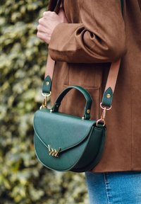 Grüne Kunstlederhandtasche mit doppeltem Griff, dünnem Riemen, goldener Hardware und einem gewölbten Klappendesign, vor einem strukturierten braunen Hintergrund.