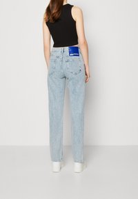 Vaqueros de denim azul claro con pierna recta, con una etiqueta rectangular azul en la cintura trasera. Textura suave y diseño minimalista.