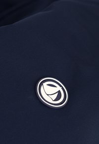 Marineblauer Stoff mit einer glatten Textur verfügt über ein rundes, weißes Logo-Patch, das darauf gesetzt ist. Das Logo hat ein geschwungenes, abstraktes Design.