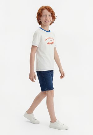 Garçon roux souriant marchant, portant un t-shirt blanc avec un texte orange, un short en jean bleu et des baskets blanches, sur un fond blanc uni.