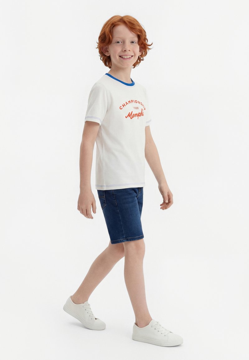 Lächelnder rothaariger Junge läuft, trägt ein weißes T-Shirt mit orangem Text, blaue Jeansshorts und weiße Sneakers vor einem schlichten weißen Hintergrund.