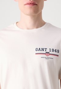 Bomulds t-shirt i off-white med korte ærmer, rund hals og frontprint af "GANT 1949" logo i marineblå og rød med et skjolddesign.