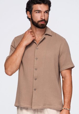 Homme avec barbe portant une chemise beige à manches courtes boutonnée, touchant le col, debout devant un fond blanc uni.