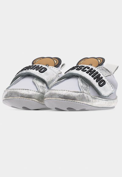 Λευκά και ασημί παπουτσάκια μωρού με ιμάντες Velcro, το γράμμα "Moschino" και ένα πρόσωπο αρκουδάκι να φαίνεται από την περιοχή της γλώσσας.
