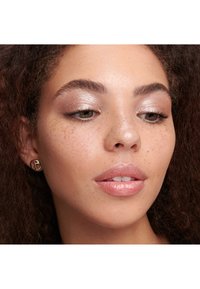 Close-up van een persoon met krullend bruin haar, die gouden ronde oorbellen draagt en een lichte, glinsterende make-up heeft. De huid heeft sproeten en de lippen zijn glanzend.