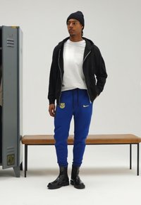 Black zip-up jacket, white t-shirt, blue sweatpants with logo, and black ankle boots; set against a neutral background with a bench.
