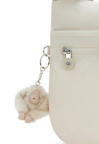 Beige skuldertaske med en glat tekstur, udstyret med lynlåslukning og en blød abe-nøglering vedhæng. Enkel, funktionel design.
