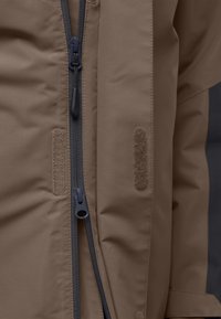 Bruine geïsoleerde jas met een centrale rits, klittenband patches en een gladde, gestructureerde stof. De manchetten hebben verstelbare banden voor een veilige pasvorm.