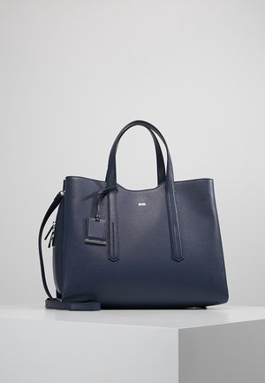 Grand sac fourre-tout en cuir bleu marine avec doubles poignées supérieures, bandoulière amovible, étiquette en cuir suspendue à l'avant, et détail subtil de logo argenté sur fond neutre.