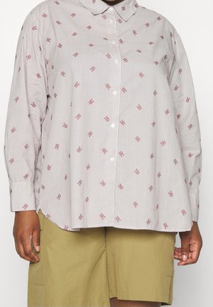 Person iført en løs, langærmet skjorte med krave og små røde mønstre, kombineret med olivengrønne shorts.