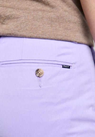 Calças lilás com uma textura suave apresentam um bolso frontal, um fecho com botão dourado e um rótulo preto "Polo" perto do bolso.