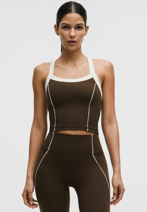 Frau mit dunklen Haaren im hohen Pferdeschwanz trägt ein dunkelbraunes, eng anliegendes Crop-Top und passende Leggings mit weißen Kanten und Nähten.