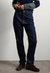 Jeans de color azul oscuro con un diseño de pierna recta, que cuentan con un cierre frontal de botón, detalles de costura clara y bolsillos. Llevados con zapatos marrones.