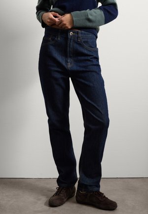 Jeans bleu foncé avec une coupe droite, dotés d'une fermeture à bouton à l'avant, de détails de couture clairs et de poches. Portés avec des chaussures brunes.